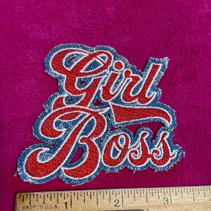 May include: An embroidered patch featuring the red text Girl Boss with a blue border. The patch is positioned above a wooden ruler that shows the item is approximately 3.5 inches wide. The lettering is in a cursive script style with a red fill.