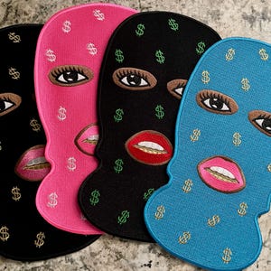 May include: Four embroidered ski masks in black, pink, black, and blue. Each mask features embroidered eyes, lips, and dollar signs. The lips are pink or red, and the eyes are brown with long lashes. The dollar signs are gold or green.