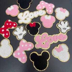 May include: Assortment of embroidered patches in various shapes and colors. Includes Mickey Mouse heads, bows, and hand shapes, along with the word "Love". Patches feature pink, white, red, and black colors with gold trim. Ideal for crafting and personalization.