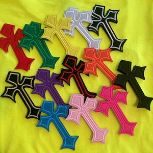 May include: A collection of embroidered cross patches in various colors including red, green, yellow, white, black, orange, purple, blue, pink, and magenta, each featuring a distinct border and a stylized, pointed shape arranged on a yellow fabric.