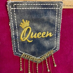 May include: A dark blue denim pocket patch featuring the embroidered yellow word Queen with a crown icon above it. The patch has white stitching along the edges and a fringe of dangling clear rhinestones at the bottom, measuring 4 inches wide.