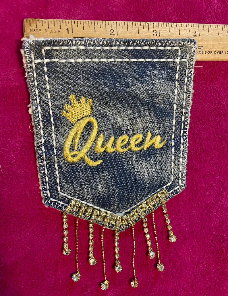 May include: A dark blue denim pocket patch featuring the embroidered yellow word Queen with a crown icon above it. The patch has white stitching along the edges and a fringe of dangling clear rhinestones at the bottom, measuring 4 inches wide.