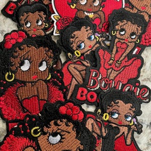 May include: A collection of embroidered patches featuring the cartoon character Betty Boop in various poses. The patches showcase Betty in red dresses and outfits, with black hair, and gold jewelry. The patches are on a light-colored surface.