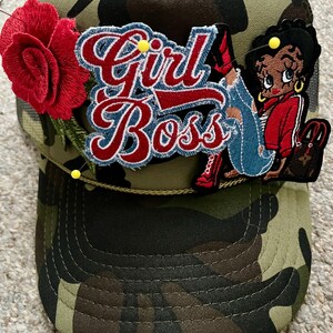 May include: A camouflage print trucker hat featuring a large red embroidered rose, a blue and red embroidered patch with the text Girl Boss, and an embroidered cartoon character patch. The hat is accented with small yellow sewing pins.