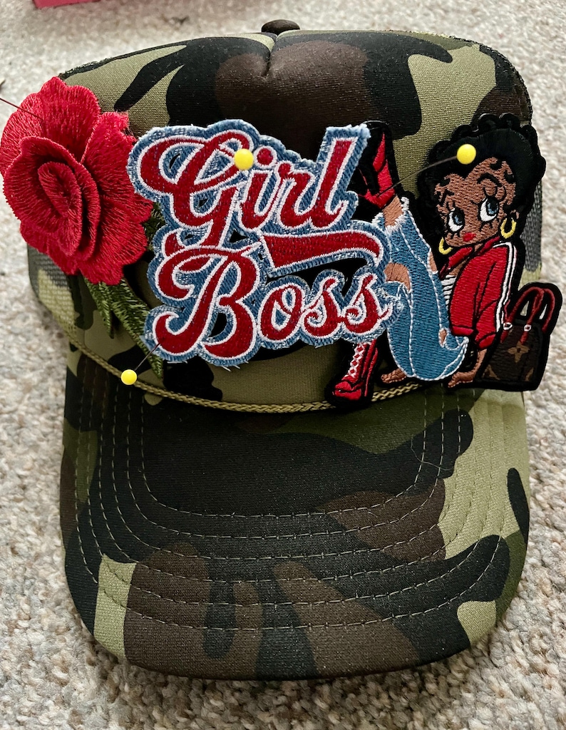 May include: A camouflage print trucker hat featuring a large red embroidered rose, a blue and red embroidered patch with the text Girl Boss, and an embroidered cartoon character patch. The hat is accented with small yellow sewing pins.