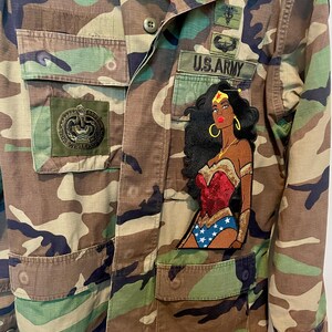 May include: A camouflage print military-style jacket featuring a large embroidered patch of a superhero character with dark hair, a red bodice, and blue star-patterned bottoms. The jacket includes a U.S. ARMY patch and other military-style insignia.