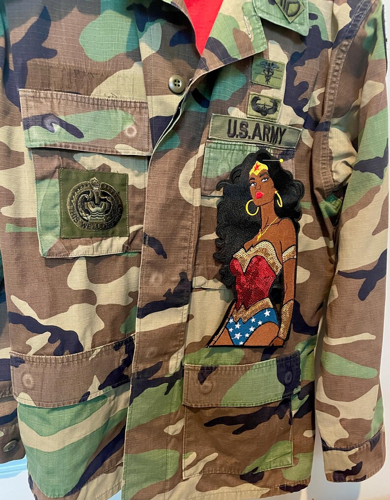 May include: A camouflage print military-style jacket featuring a large embroidered patch of a superhero character with dark hair, a red bodice, and blue star-patterned bottoms. The jacket includes a U.S. ARMY patch and other military-style insignia.