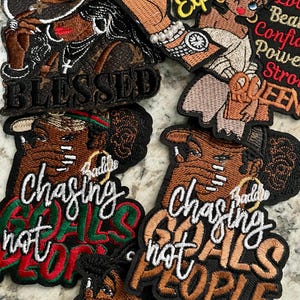 May include: A collection of embroidered patches with various designs and text. One patch reads "BLESSED" in white lettering. Other patches feature illustrations of women and phrases like "Chasing Goals Not People" and "Smell Expensive".