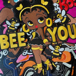 May include: An embroidered patch featuring a cartoon character with dark skin, black hair, and yellow hair accessories, wearing a black outfit. The text BEE YOU is embroidered in yellow. The design includes bees and a motorcycle on a pink background.