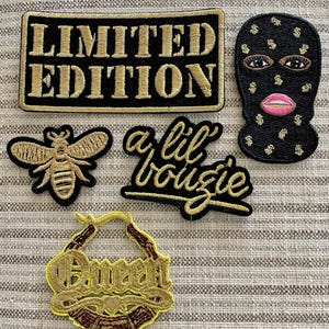 May include: Five embroidered iron-on patches with gold thread on a black background. The patches include the words "Limited Edition", "a lil boujie", a bee, a gold hoop earring with the word "Queen" inside, and a black ski mask with gold dollar signs.