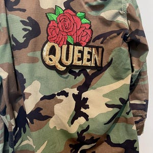 May include: A camouflage print jacket featuring a large embroidered patch on the back. The patch displays three red roses with green leaves above the word QUEEN, which is written in gold-colored sequins on a black background.