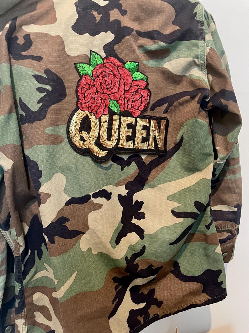 May include: A camouflage print jacket featuring a large embroidered patch on the back. The patch displays three red roses with green leaves above the word QUEEN, which is written in gold-colored sequins on a black background.