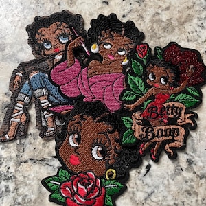 May include: A collection of embroidered patches featuring the cartoon character Betty Boop. The patches showcase Betty in various poses, with vibrant colors and detailed stitching. One patch includes the text "Betty Boop" on a banner, and another features a red rose.