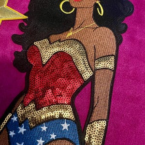 May include: An embroidered patch featuring a stylized figure with dark skin, long black hair, and gold hoop earrings. The figure wears a red and gold sequined bodice, a blue skirt with white stars, gold arm cuffs, and a gold tiara with a red star.