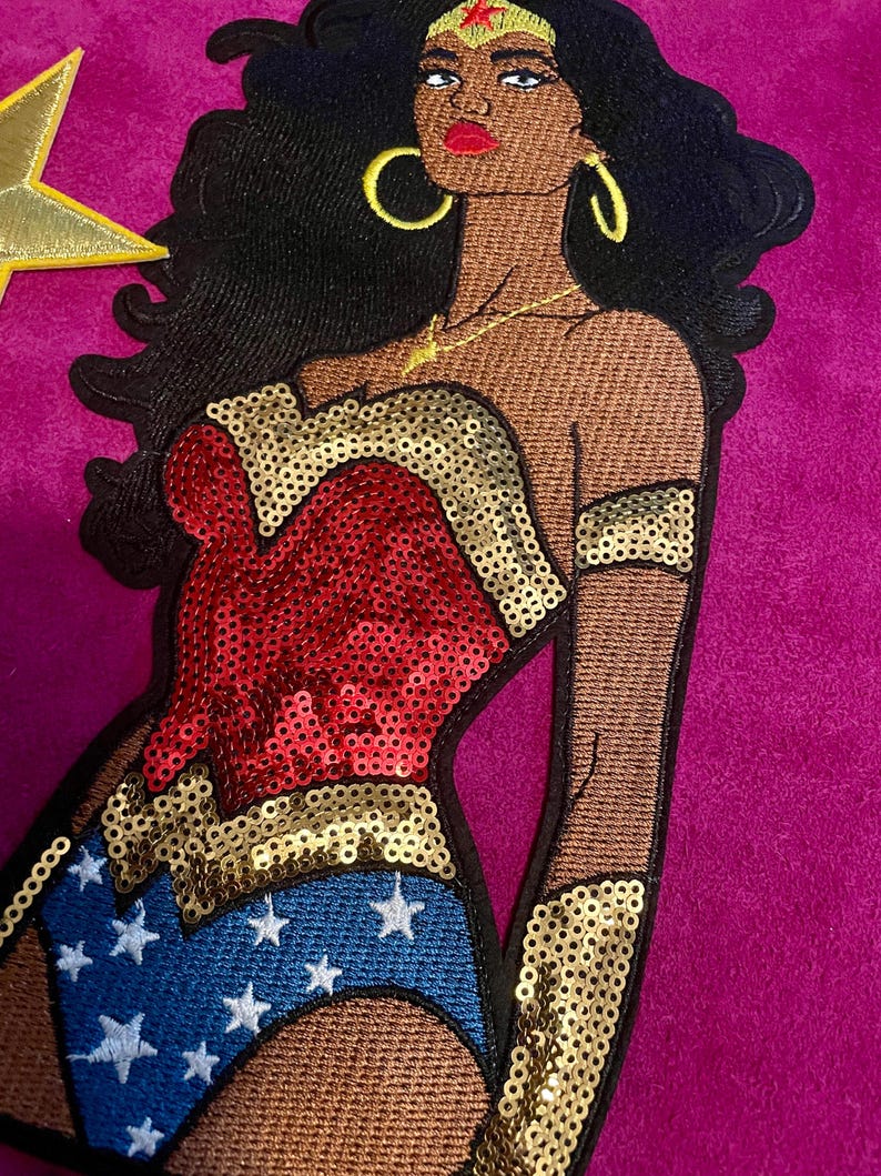 May include: An embroidered patch featuring a stylized figure with dark skin, long black hair, and gold hoop earrings. The figure wears a red and gold sequined bodice, a blue skirt with white stars, gold arm cuffs, and a gold tiara with a red star.