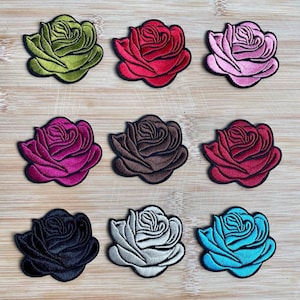 May include: Nine embroidered rose patches in various colors, including green, red, pink, magenta, brown, burgundy, black, white, and teal. Each patch features a detailed rose design with a black border, suitable for fabric embellishment.