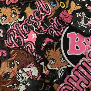 May include: A collection of embroidered patches with various designs and text. The patches feature cartoon characters, the word "Baddie", and the word "Blessed" in pink and black. The patches are likely for clothing or accessories.