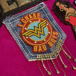 May include: A denim patch featuring a circular emblem with a yellow Wonder Woman logo on a light blue background, surrounded by a red border with the text I CHASE BAD BOYS. The patch has dangling silver-toned chains with clear rhinestones.
