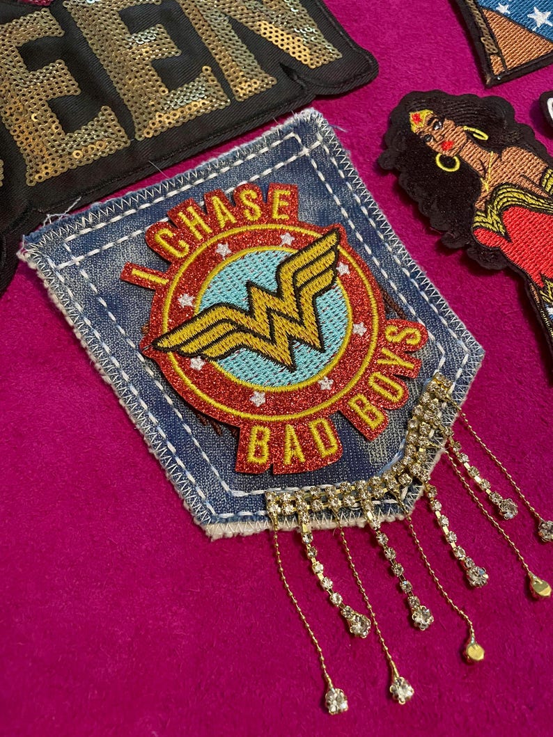 May include: A denim patch featuring a circular emblem with a yellow Wonder Woman logo on a light blue background, surrounded by a red border with the text I CHASE BAD BOYS. The patch has dangling silver-toned chains with clear rhinestones.