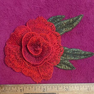 May include: A red rose embroidered patch with dark green leaves, positioned above a wooden ruler that shows the flower measures approximately 3.5 inches in width. The embroidery features dense, layered stitching to create a floral shape.
