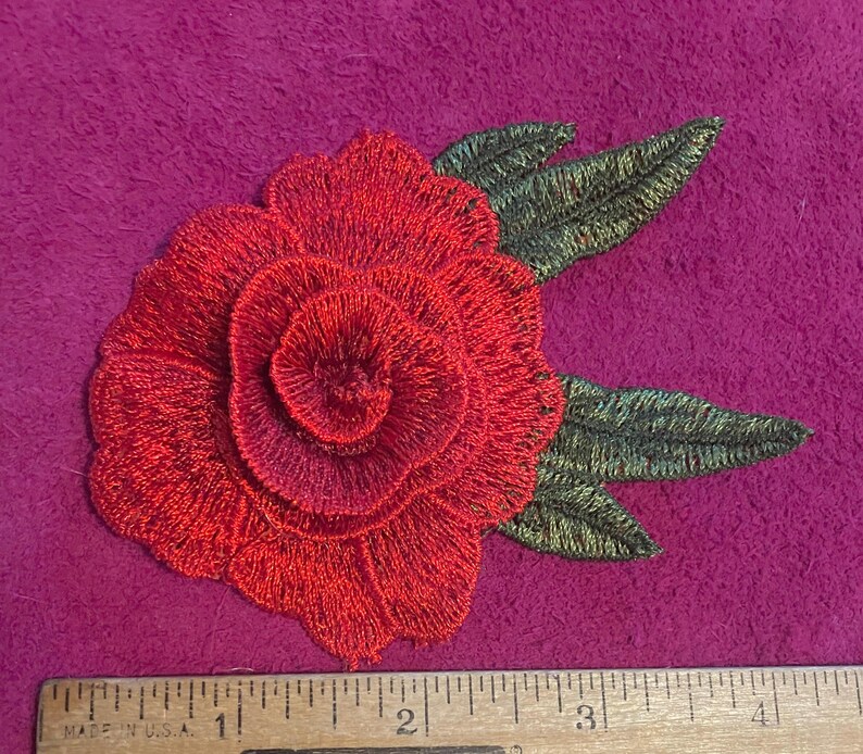 May include: A red rose embroidered patch with dark green leaves, positioned above a wooden ruler that shows the flower measures approximately 3.5 inches in width. The embroidery features dense, layered stitching to create a floral shape.