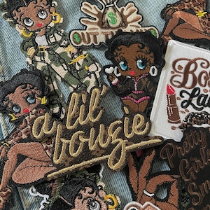 May include: A collection of embroidered patches featuring cartoon characters in various poses and outfits. Some patches include text like "I Got It Out", "Lil Bougie", and "Boss Lady". The patches are in shades of brown, black, green, and pink.