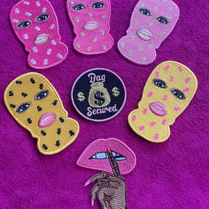 May include: A collection of embroidered patches featuring four balaclava masks in pink and yellow with dollar sign patterns, a circular patch with a money bag graphic and the text Bag Secured, and a patch of a hand holding a pair of pink lips.