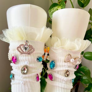 May include: Pair of white, ribbed socks with a cream-colored tulle ruffle at the top. Embellished with a crown, bow, and various colorful jewels and faux pearls. The socks are displayed on white cylindrical forms.