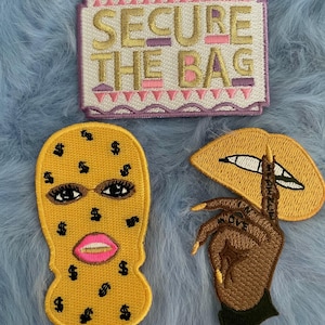 May include: Three embroidered iron-on patches. One patch is yellow with dollar signs and a pink mouth, another is a brown hand with long gold nails, and the third is white with pink and purple accents and the text "SECURE THE BAG".