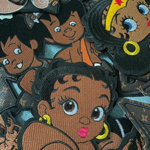 May include: A collection of embroidered patches featuring cartoon characters. The patches showcase a variety of designs, including a character with a star headband, and a character with a yellow earring. The patches are in various shades of brown, black, and yellow.
