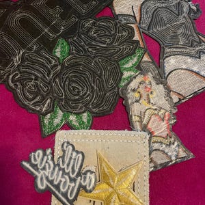 May include: A collection of embroidered patches including a black patch with the word QUEEN and black roses, two patches depicting a figure with long hair and gold jewelry, a small black patch with white script text, and a gold star on a beige fabric.