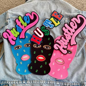 May include: Three colorful embroidered patches featuring balaclava designs with dollar signs and the word "Hustle". The patches are in blue, black, and pink, with the word "Hustle" in pink, green, and blue. The patches are on a denim jacket.