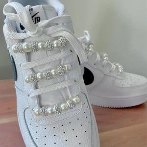 May include: White sneakers with pearl and rhinestone accents on the laces. The shoes have a classic design with a black Nike swoosh logo on the side. The laces are white, and the shoes have a perforated toe box.