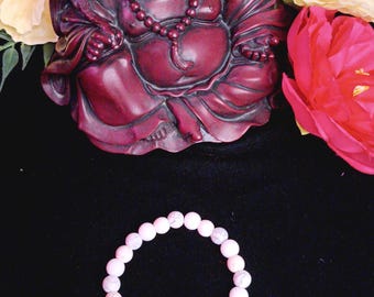 May include: A white beaded bracelet with a rainbow of colored stones. The bracelet is lying on a black surface in front of a red statue of a laughing Buddha.