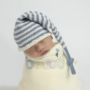 Puede incluir: Un bebé recién nacido con un gorro de punto azul y blanco a rayas con una cola larga, durmiendo en una manta de arrullo blanca con la palabra "bebé" bordada.