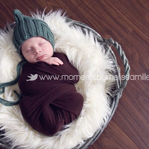 Może przedstawiać: Noworodek owinięty w ciemnobrązowy kocyk, w zielonej dzianinowej czapce z nausznikami, spoczywający w wiklinowym koszu wyłożonym białym sztucznym futrem. Kosz stoi na ciemnej drewnianej powierzchni. Tekst na zdjęciu: www.momentsbymelissamiller.com.