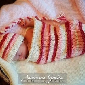 May include: A newborn baby is swaddled in a pink and white striped knitted blanket and wearing a matching knitted hat. The baby is sleeping peacefully.