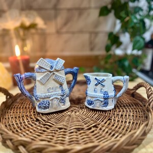May include: A set of two blue and white ceramic sugar and creamer containers shaped like windmills. The containers are sitting on a brown wicker tray.