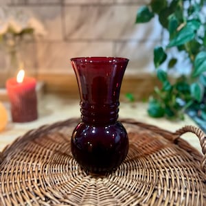 May include: A small, dark red glass vase with a rounded base and a flared top. The vase is sitting on a woven wicker tray.