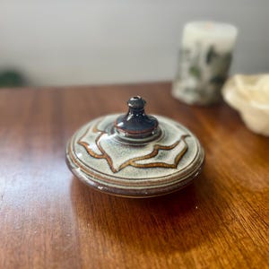 May include: A small, white ceramic incense burner with brown and gold accents. The burner has a round base and a tall, narrow spout.