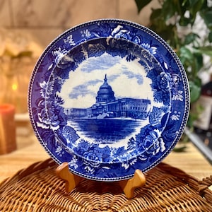 May include: A round ceramic plate featuring a blue and white transferware pattern. The center depicts the United States Capitol building, while the wide border is decorated with a dense arrangement of detailed blue roses and leafy floral vines.