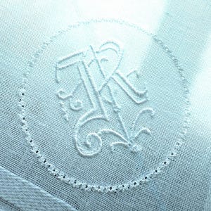 Könnte beinhalten: Nahaufnahme eines hellblauen Taschentuchs mit einem gestickten Monogramm in einem kreisförmigen Rahmen. Das Monogramm zeigt verschlungene Buchstaben, der Rand ist mit kleinen, gleichmäßig verteilten Löchern versehen.