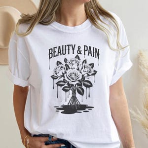 以下が含まれることがあります： 「BEAUTY & PAIN」の文字と、バラの花束の白黒イラストがプリントされた白いTシャツ。 バラは茎と葉で束ねられ、滴るようなアクセントが施されています。