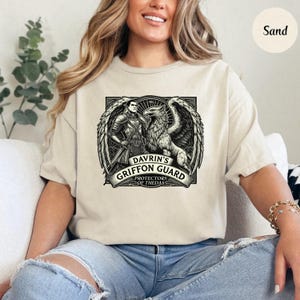 May include: A beige t-shirt with a black and white graphic print of a knight and a griffin. The graphic includes the text "Davrin's Griffon Guard, Protectors of Thedas".