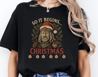 Peut inclure: T-shirt noir avec un motif festif. Le design présente un homme portant un bonnet de Père Noël et une armure, entouré de houx et d'ornements. Le texte dit "SO IT BEGINS... CHRISTMAS".