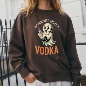 Könnte beinhalten: Ein braunes Sweatshirt mit einer Grafik eines Cartoon-Geistes, der eine Wodkaflasche hält. Der Text "MY FAVORITE SPIRIT IS VODKA" ist über und unter der Geisterillustration aufgedruckt. Das Sweatshirt ist einfarbig.