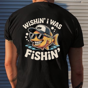 May include: Black t-shirt featuring a cartoon fish wearing sunglasses and a hat. The fish is yellow with orange and brown accents, and the text "WISHIN' I WAS FISHIN'" is above and below the fish in white lettering. The shirt is made of a soft material.