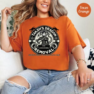 May include: A woman wearing an orange t-shirt with a black and white graphic of a dragon and the text "Taash's Dragon Removal, The Veilguard, Serving Thedas, One Dragon At A Time".