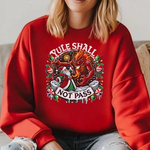 Puede incluir: Sudadera roja de Navidad con un gráfico de Papá Noel luchando contra un demonio rojo con el texto "Yule Shall Not Pass".