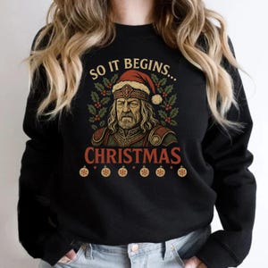 May include: Black sweatshirt with a graphic featuring a man wearing a Santa hat and armor. The text reads "SO IT BEGINS... CHRISTMAS" with decorative elements. Below the text are six ornaments.